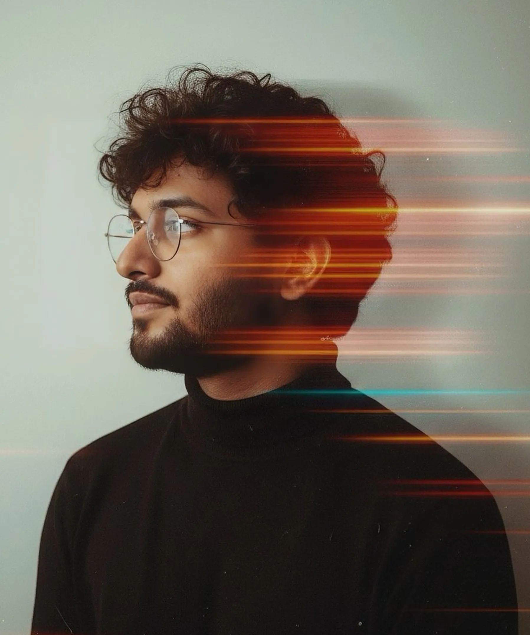 Young man with curly hair, beard, and glasses wearing a black turtleneck, with orange and blue light streaks across his profile.