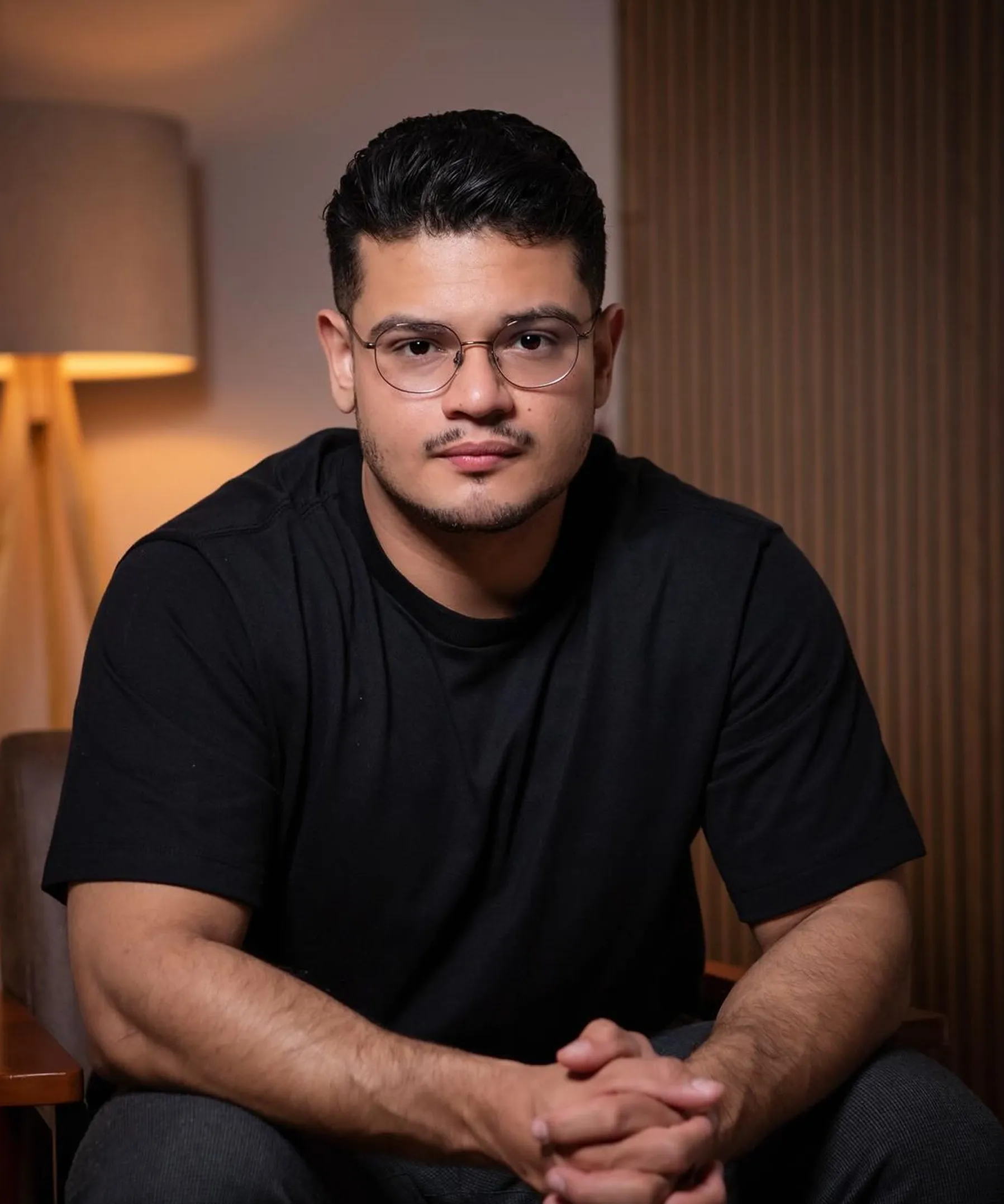 Young man with glasses and short dark hair wearing a black t-shirt, sitting with hands clasped indoors with a lamp and brown wooden panel background.