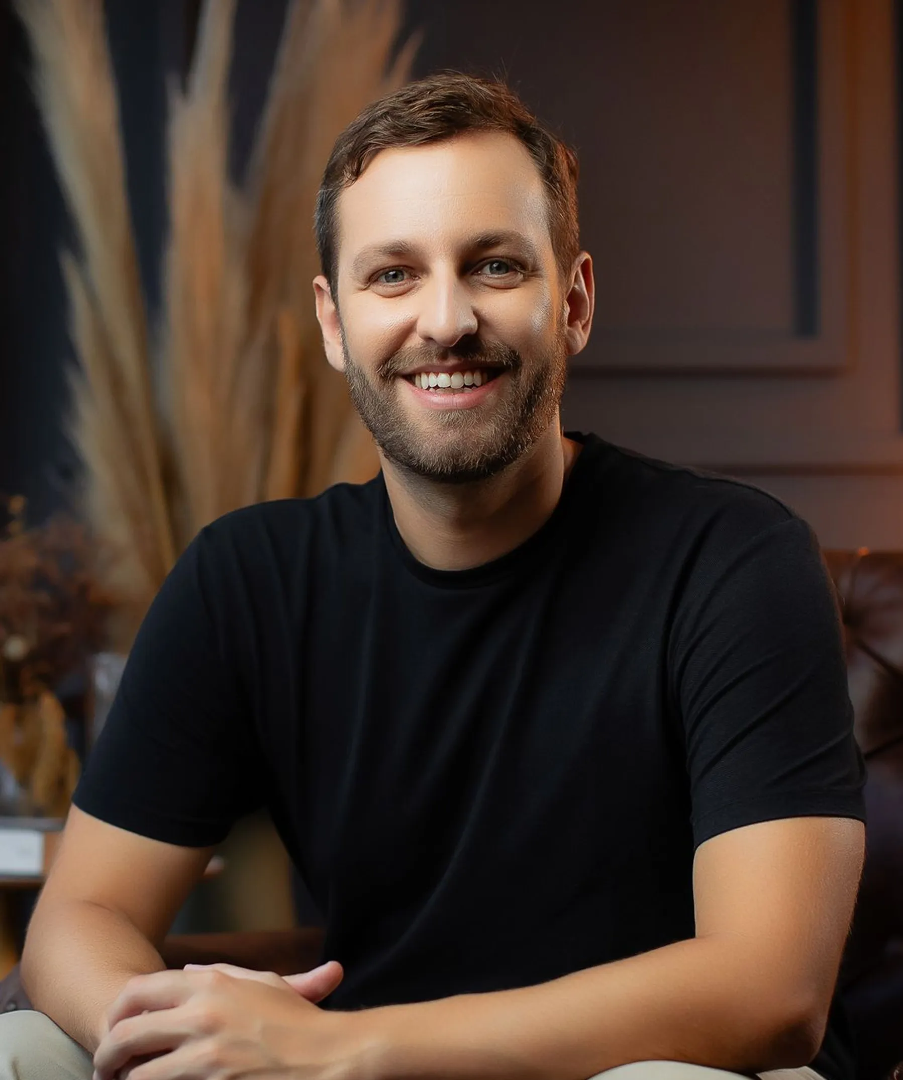 Smiling man with short brown hair and a beard wearing a black t-shirt, seated indoors with a blurred background.
