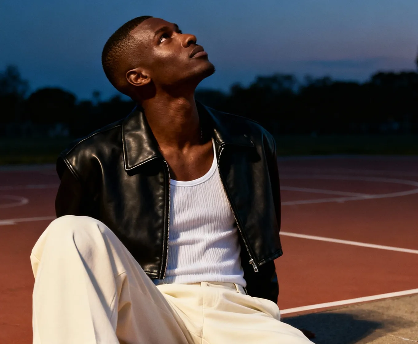 Young man in a black leather jacket and white tank top sitting on an outdoor basketball court at twilight, looking upward.